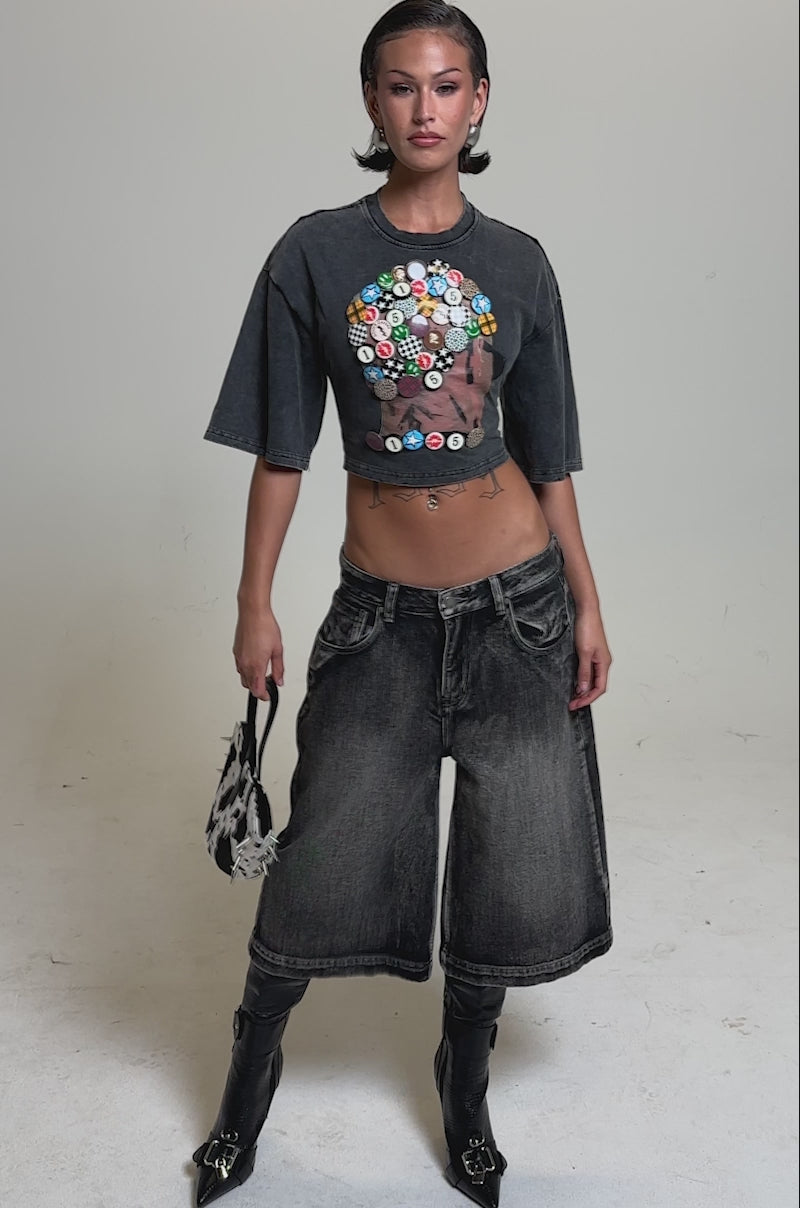 A video of a woman in the PINNED DOWN MINERAL WASHED TEE IN GREY, wide-leg faded black jeans, black high-heeled boots, and a cow-print handbag. Her hair is slicked back as she stands against a plain background.