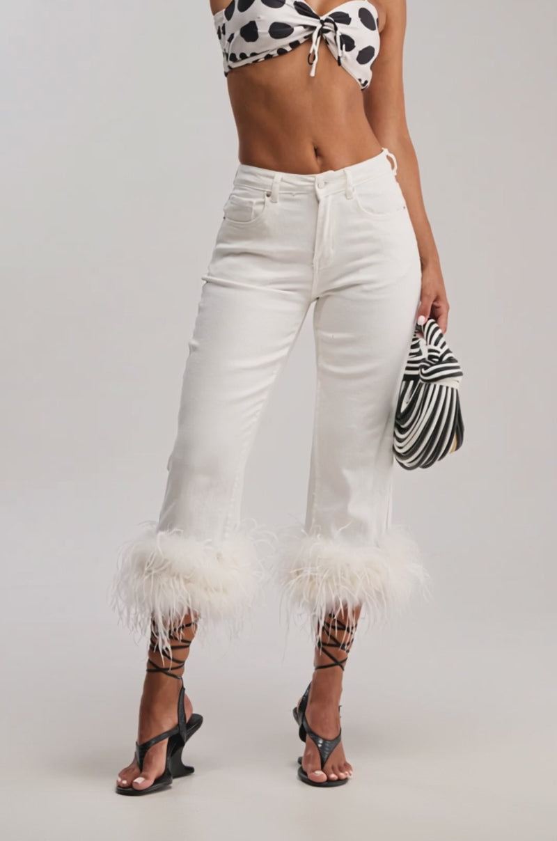 Video of woman modeling the TRIPPY LIKE ME DENIM PANT WITH FEATHER, styled with strappy black sandals and a black-and-white striped handbag. Her midriff is visible against a plain light gray background.