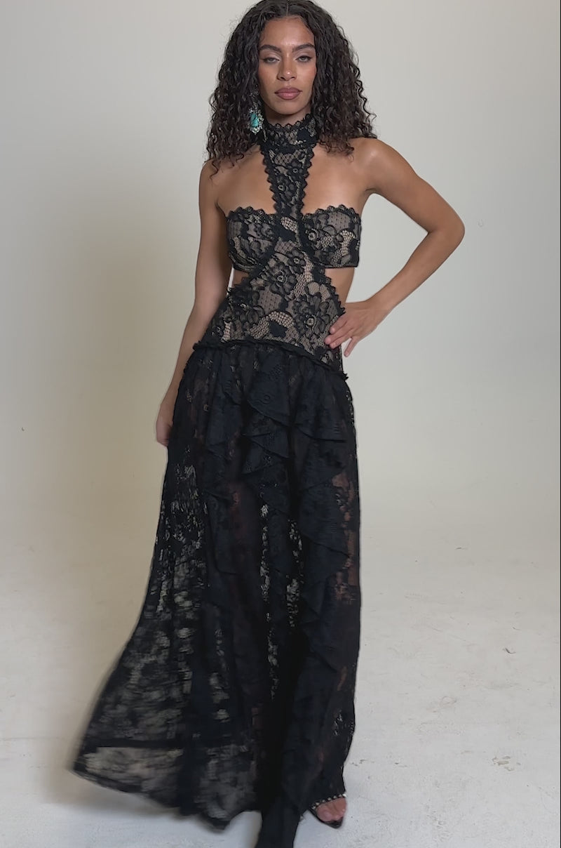 Video of woman modeling the KYLA LACE MAXI DRESS IN BLACK—a sleeveless gown with a sculpted silhouette, high neckline, sheer lace bodice, and ruffled skirt. The intricate patterns stand out against a plain, light background.
