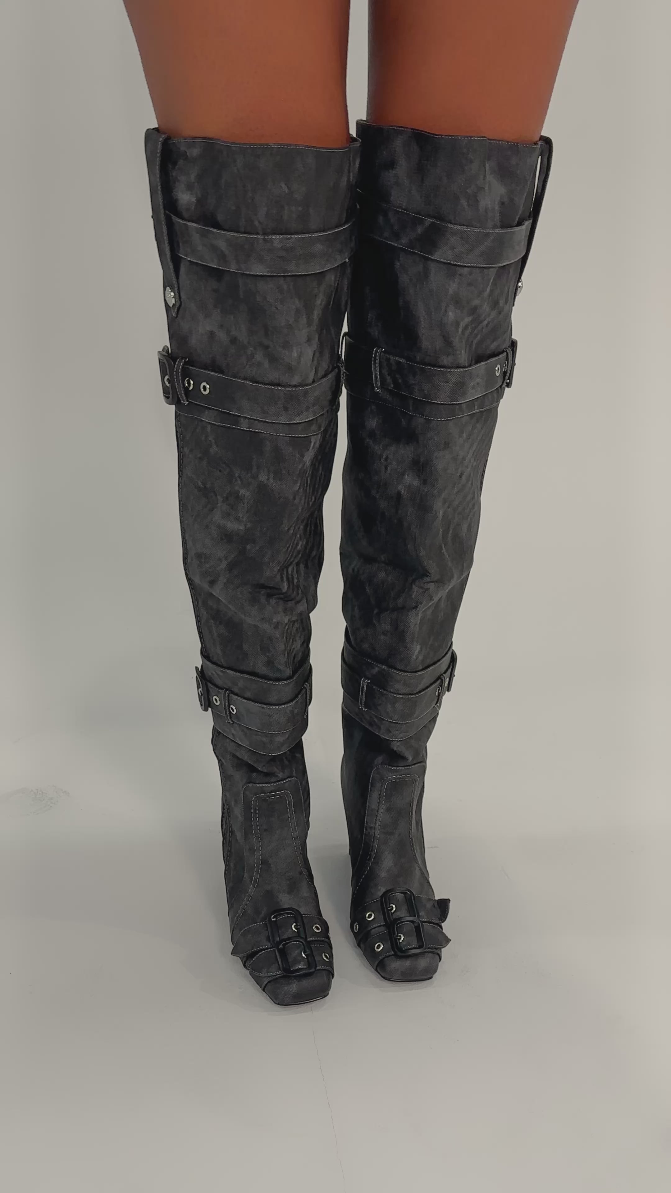 A video of a person wearing the AZALEA WANG TRILLION CHARCOAL CHUNKY THIGH HIGH BOOT, featuring black over-the-knee design with buckles and straps, standing on a plain light background to showcase its bold style.