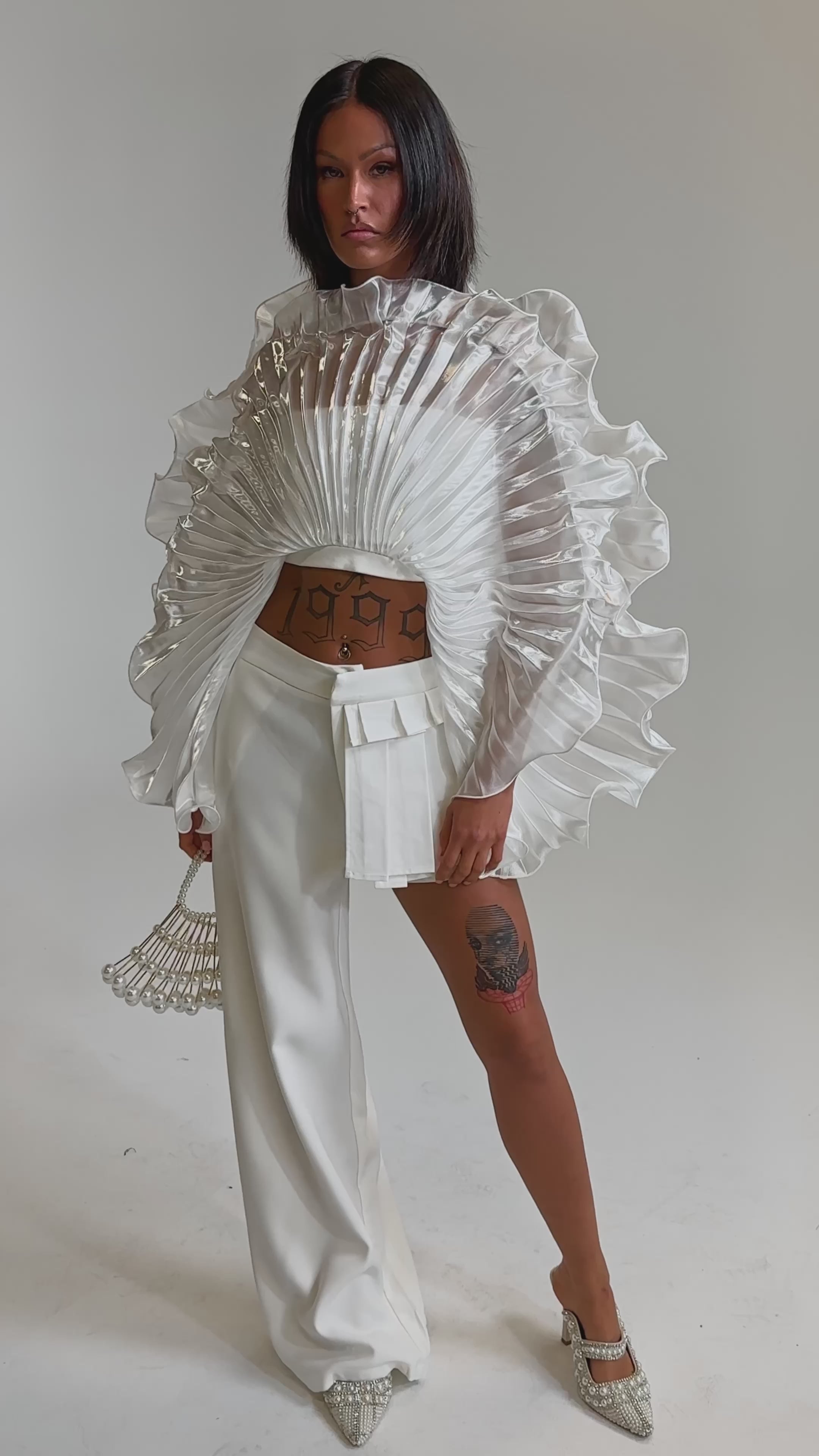 A video of a woman in a ruffled, sheer fan-shaped white top and the HOT AND COLD HYBRID PANT IN WHITE—an asymmetric design with one long and one short leg—paired with a beaded handbag, tattoos, and pointed shoes.
