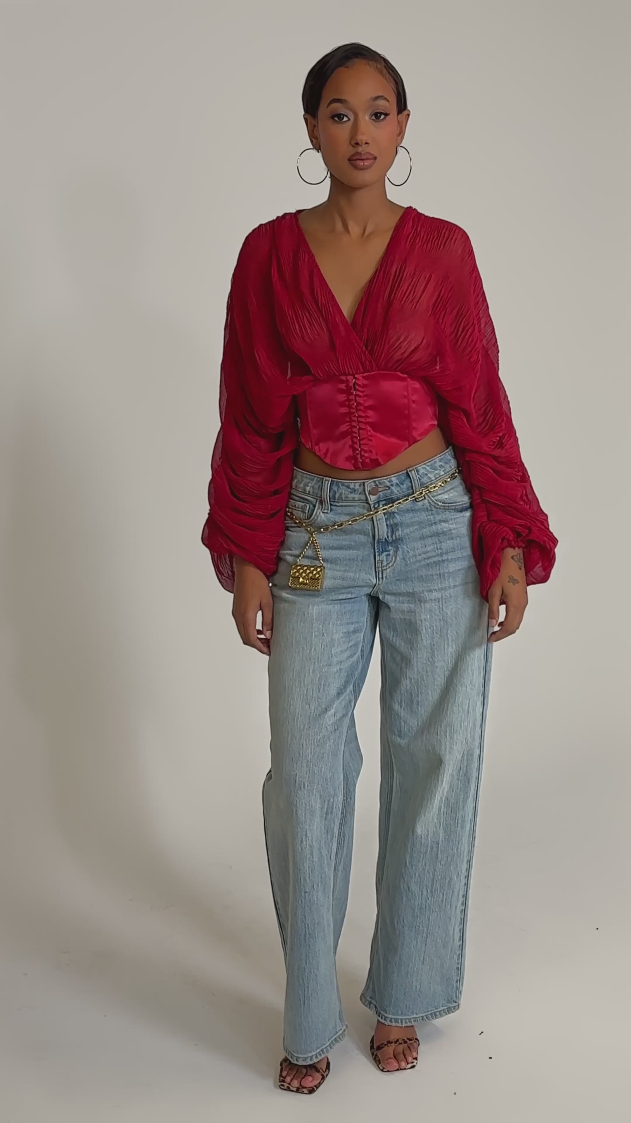 A video of a woman wearing the GIRLY PLZ BLOUSE in red chiffon with ruched balloon sleeves, paired with light blue wide-leg jeans, gold hoop earrings, layered gold belts, and open-toed heels.