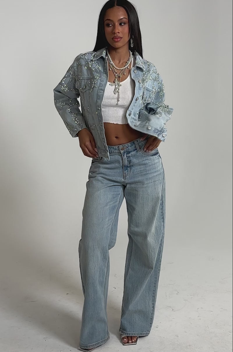 A video of a woman in the TWILIGHT ZONE RHINESTONE DENIM JACKET over a white crop top and loose light blue jeans. 