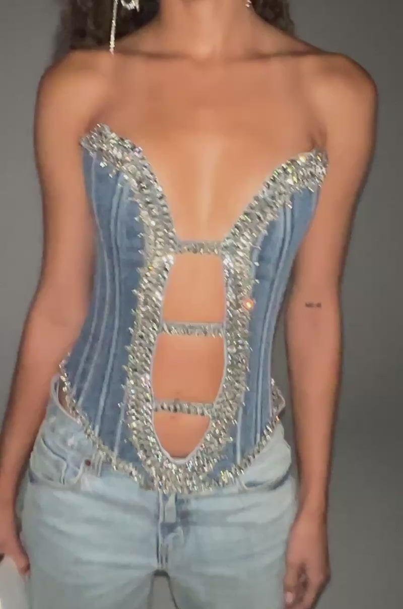 Video of woman modeling the LUX LIFE CORSET IN LIGHT BLUE DENIM, featuring rhinestones, sparkly embellishments, and cutouts. She pairs it with light blue jeans, a silver clutch, and large hoop earrings for a standout brunch look.