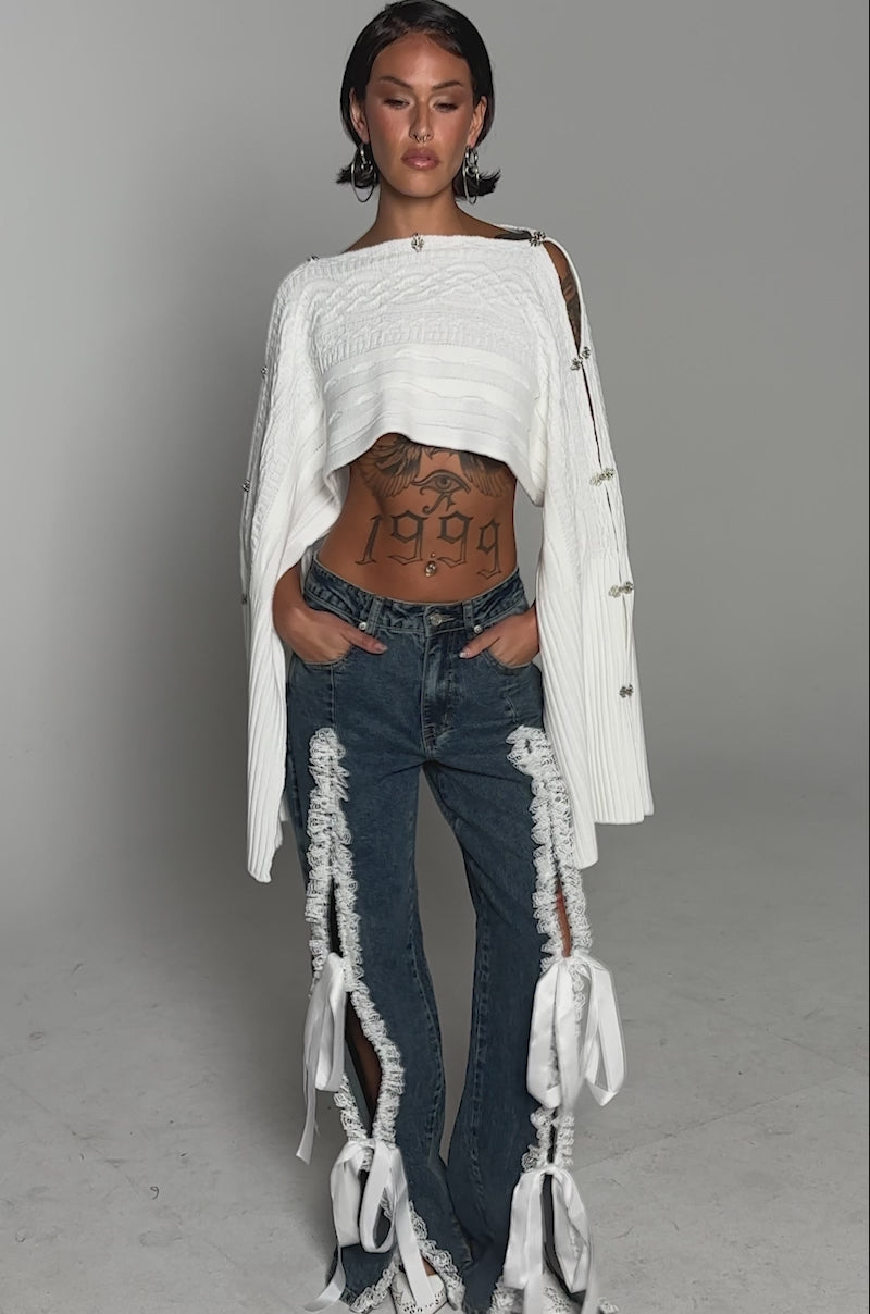 A video of a woman standing against a plain background wearing TIE ME UP RELAXED DENIM—high-rise jeans with lace trim and white bows—paired with a cropped white sweater, white shoes, hoop earrings, and short hair, revealing her midriff tattoos.