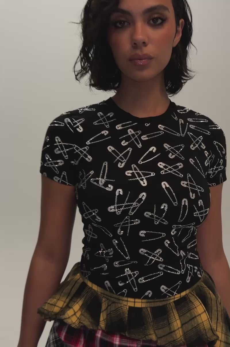 Video if a woman modeling the PROCEED WITH CAUTION RHINESTONE TEE, a streetwear-inspired black t-shirt adorned with white safety pin patterns, paired with a plaid skirt featuring yellow and red layer.