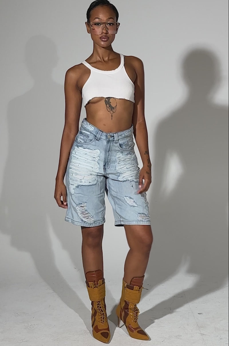 A video of a person standing against a plain background wearing a white cropped top, KIMMY DISTRESSED BERMUDA SHORTS, yellow heeled boots, and clear glasses.