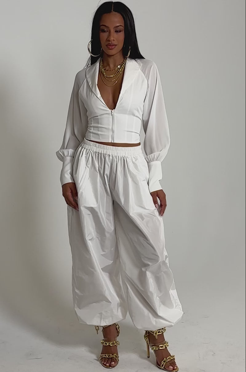 A video of a woman in the EFFORTLESS CORSET BLOUSE TOP, a chic white zip-up blouse paired with loose pants, gold chain necklaces, and gold high-heeled sandals.
