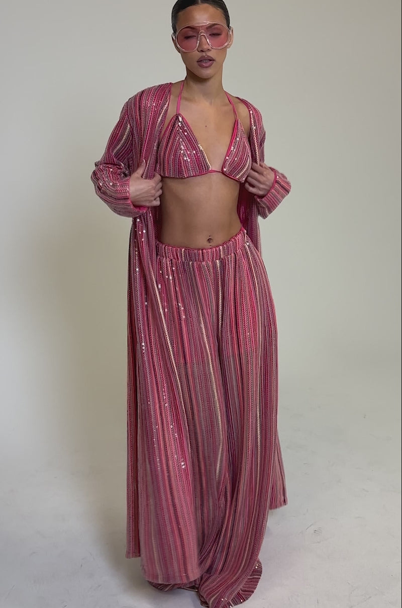 Video of woman modeling the TAYLOR SEQUIN BRALETTE, a woman styles a pink, shimmering loungewear set with wide-leg pants and a long duster. She completes her look with pink-tinted sunglasses and slicked-back hair.