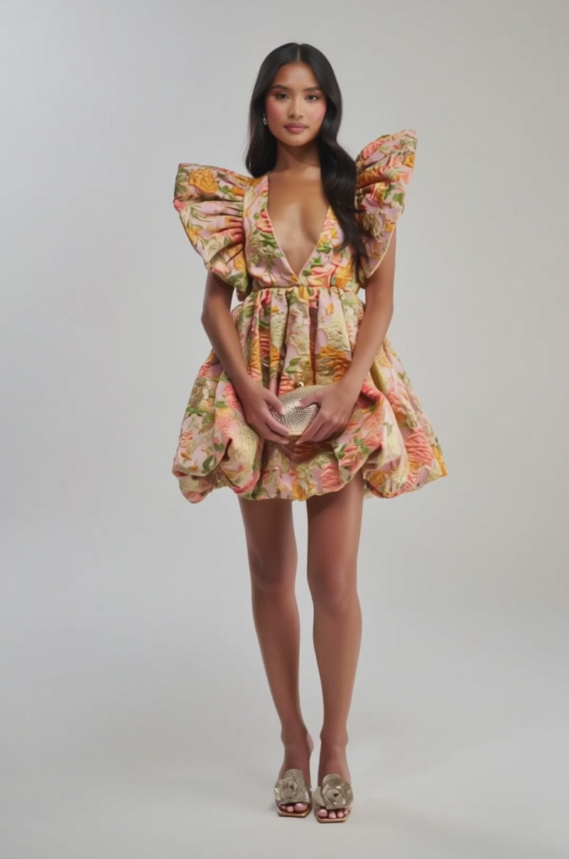 Video of woman modeling the MAIN CHARACTER ENERGY BROCADE MINI DRESS IN PEACH—a floral mini with dramatic puffed sleeves, a deep V-neckline, and pink, gold, and green tones. She carries a small handbag.