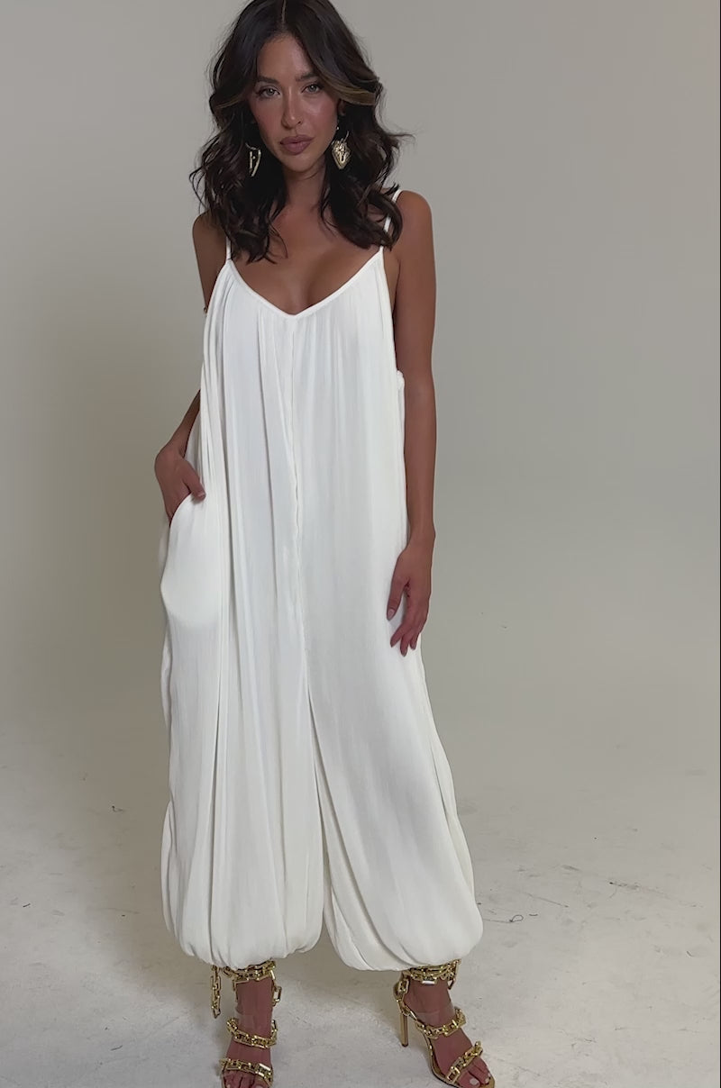 Video of woman modeling the HALO SLINKY SLEEVELESS JUMPSUIT with gathered ankles, gold high-heeled sandals, and hoop earrings 