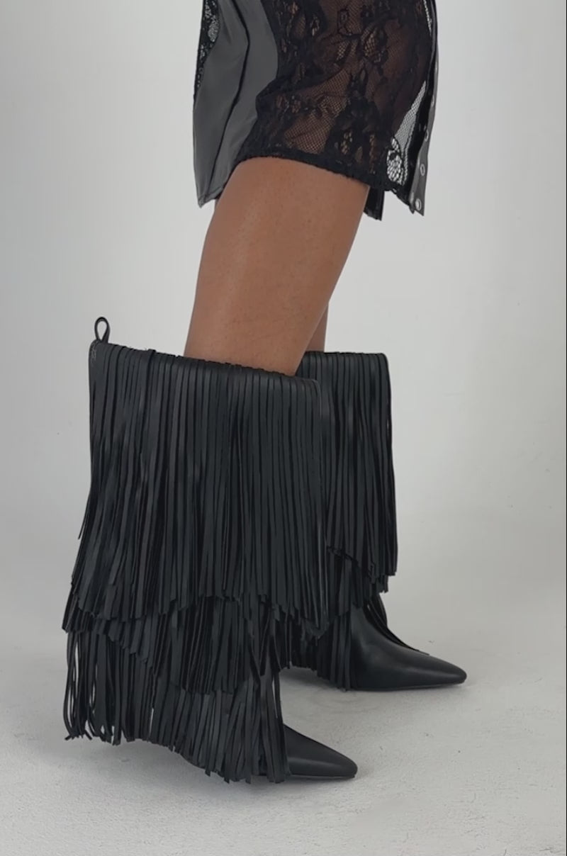 Video of a woman modeling the Wearing the AZALEA WANG FIFER BLACK FRINGE STILETTO BOOTIE, a person pairs knee-high black fringe stiletto booties with a black lace slit skirt and carries a textured black handbag against a plain light-colored background.