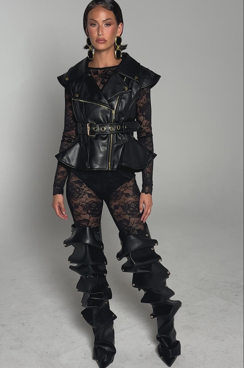 A video of a woman in the PEP IN MY STEP LEATHER VEST—a sleeveless black faux leather vest with an adjustable waist-belt—paired with a sheer lace bodysuit and tall black boots, against a light gray background.