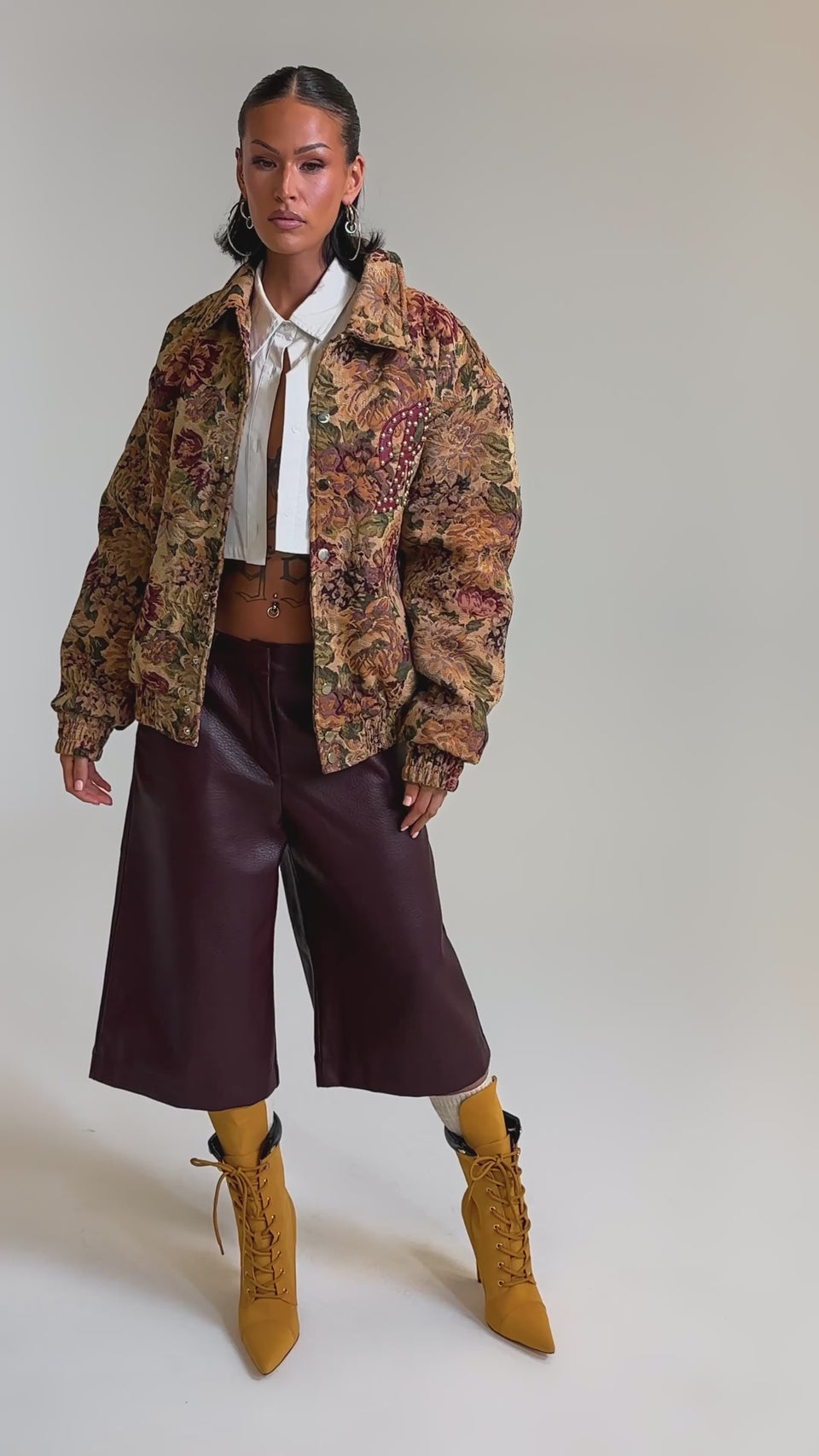 A video of a person in the SLIDE ON ME TAPESTRY BOMBER, an unbuttoned white shirt, high-waisted brown culottes, and mustard yellow heeled boots against a plain light background.