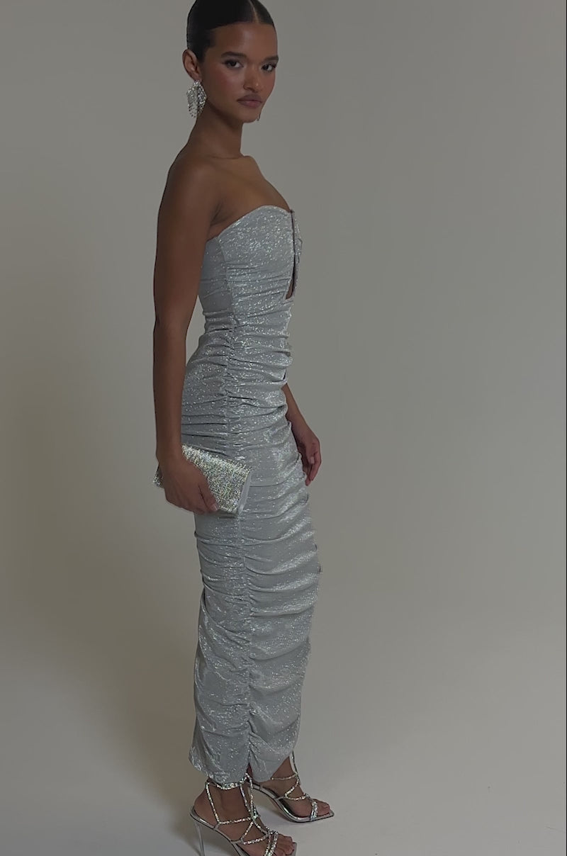 Video of woman modeling the CANT GET ENOUGH SLINKY RUCHED MAXI DRESS, featuring a strapless, glitter design with ruching and a front cutout. She completes the look with strappy heels, statement earrings, and a silver clutch against a gray backdrop.