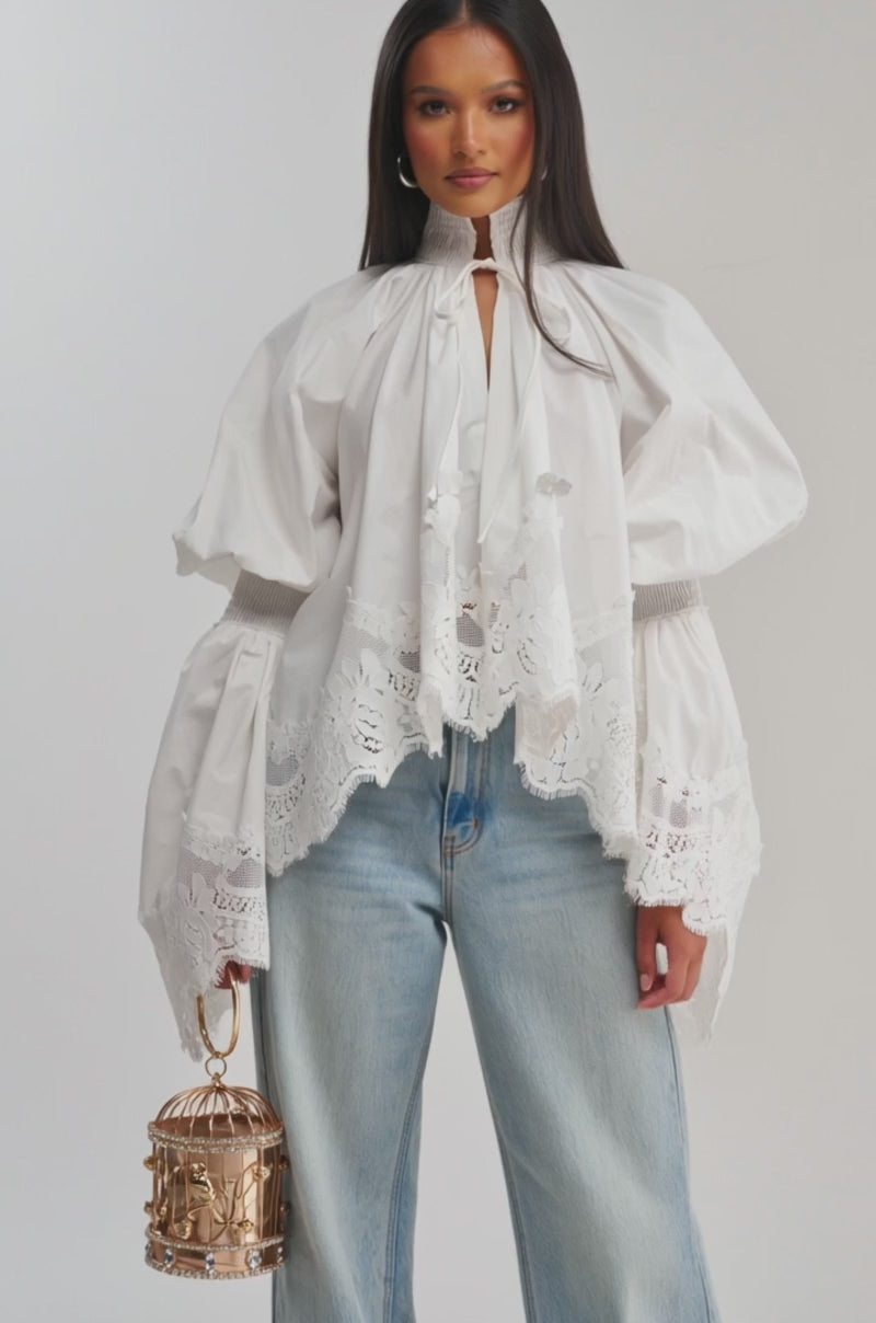 Video of woman modeling the MARIA LACE TRIM POPLIN BLOUSE with billowy sleeves and lace accents, paired with light blue jeans. She holds a small round handbag and stands against a simple light background.