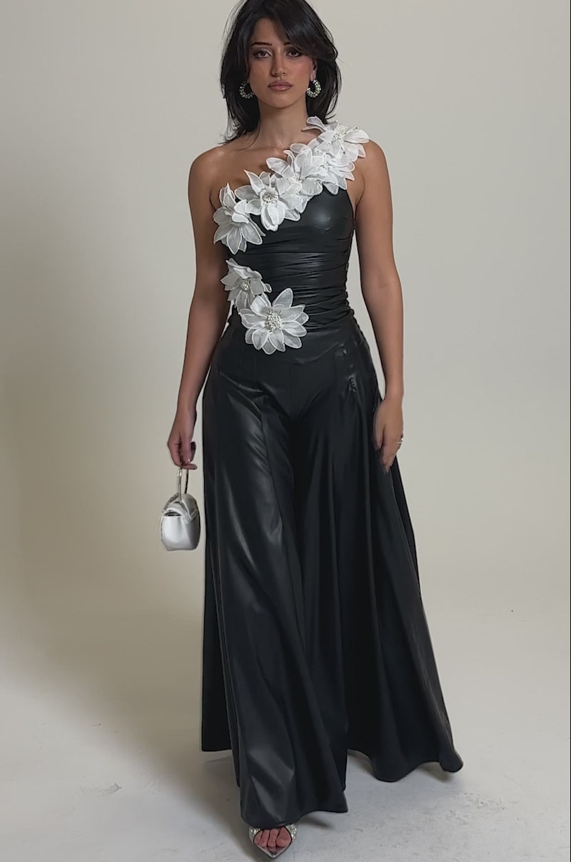 A video of a woman wearing the FLORAL BABY ONE SHOULDER BLOUSE, a black one-shoulder jumpsuit with wide-leg pants and bold white floral appliqués on the top and shoulder, set against a plain light gray background.