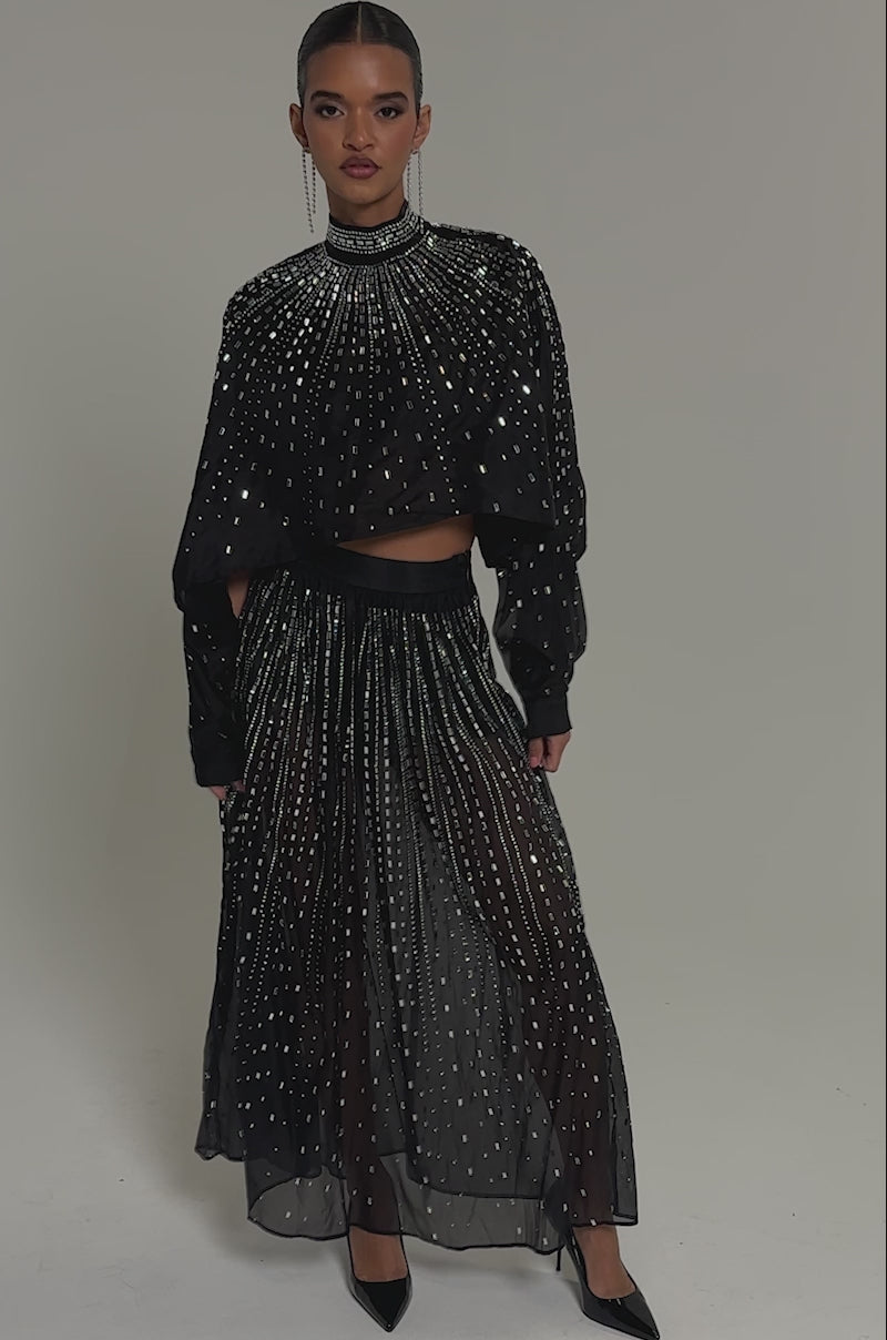 Video of woman modeling the DANCING QUEEN RHINESTONE MESH MAXI SKIRT, a black sheer outfit with sparkling rhinestones, styled with a loose long-sleeve top over black shorts and finished with black pointed heels.