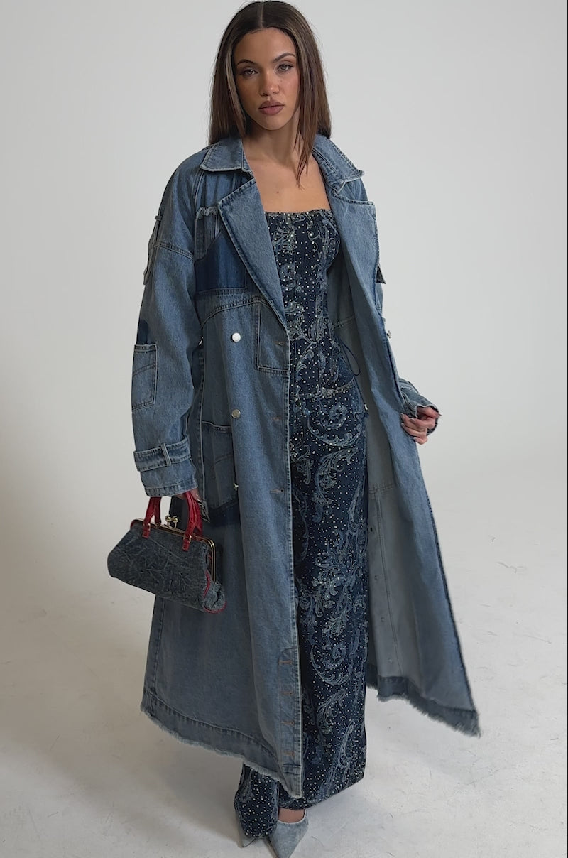 Video of woman modeling the RILEY PATCHWORK DENIM TRENCH, a vintage-wash longline coat with silver buttons, styled over blue patterned pants. She carries a small handbag and pointed shoes, posing against a plain white background.