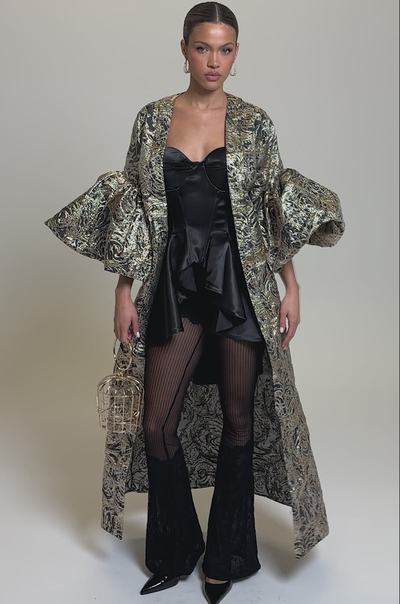 Video of woman modeling the METALLIC PUFF SLEEVE BROCADE DUSTER IN SILVER over a black corset dress, sheer striped tights, and pointed black boots, accessorizing with a small gold handbag.