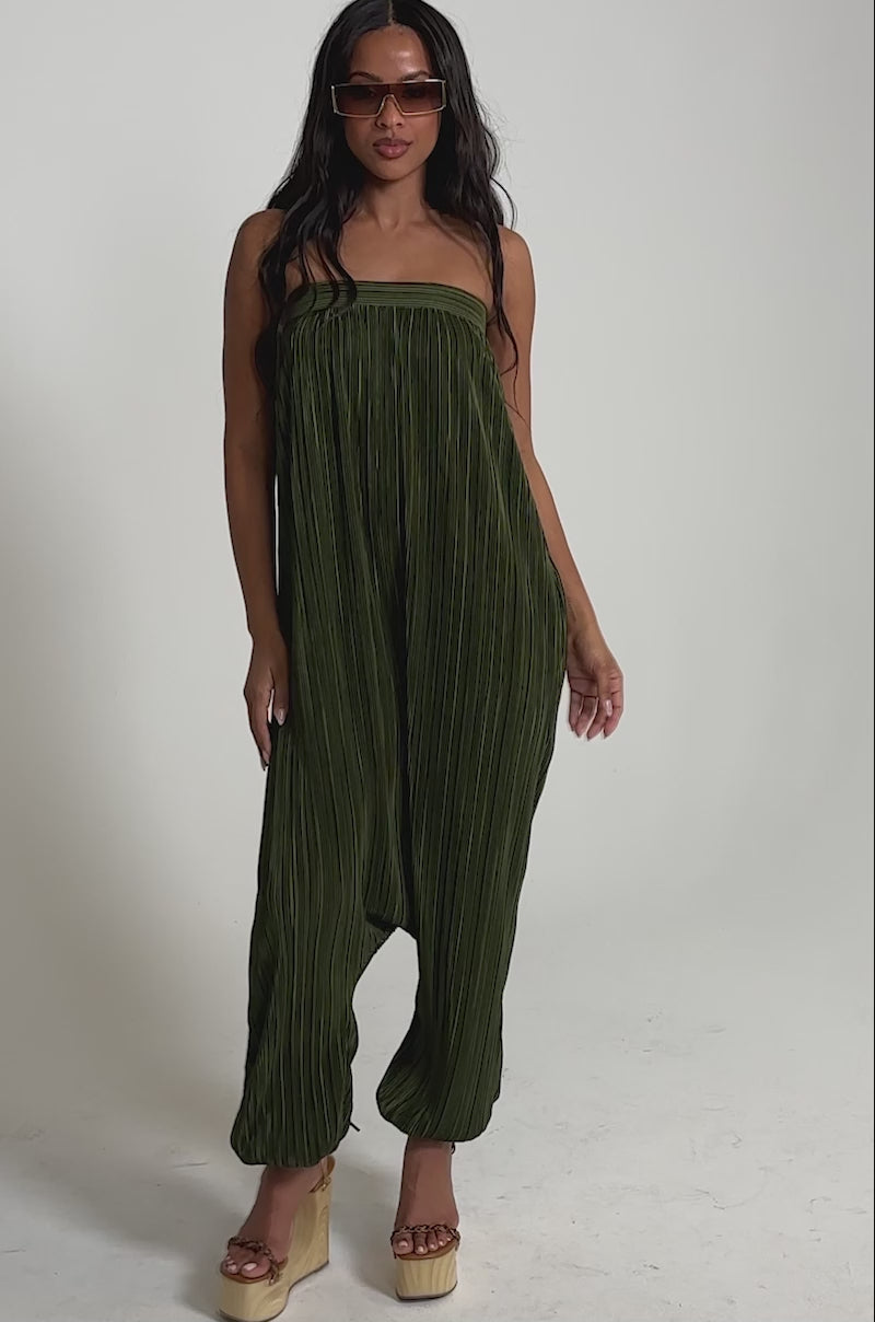 Video of woman modeling the FROM THE CITY DROP CROTCH JUMPSUIT IN OLIVE—an olive green, strapless jumpsuit with pleated harem-style pants—styled with platform sandals and sunglasses, ideal for resortwear