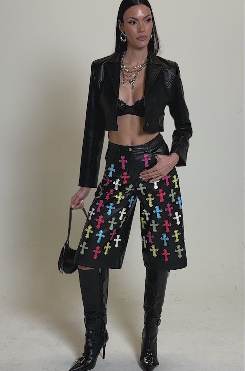 A video of a woman in the REAL ONE FAUX LEATHER BLAZER over a black lace bralette and colorful cross-patterned pants, accessorized with layered necklaces, knee-high black boots, and a chic black bag.