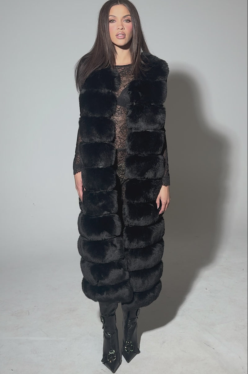 Video of woman modeling the JENNY LIN FAUX FUR MAXI VEST IN BLACK layered over a sheer lace dress and paired with sleek black knee-high boots.