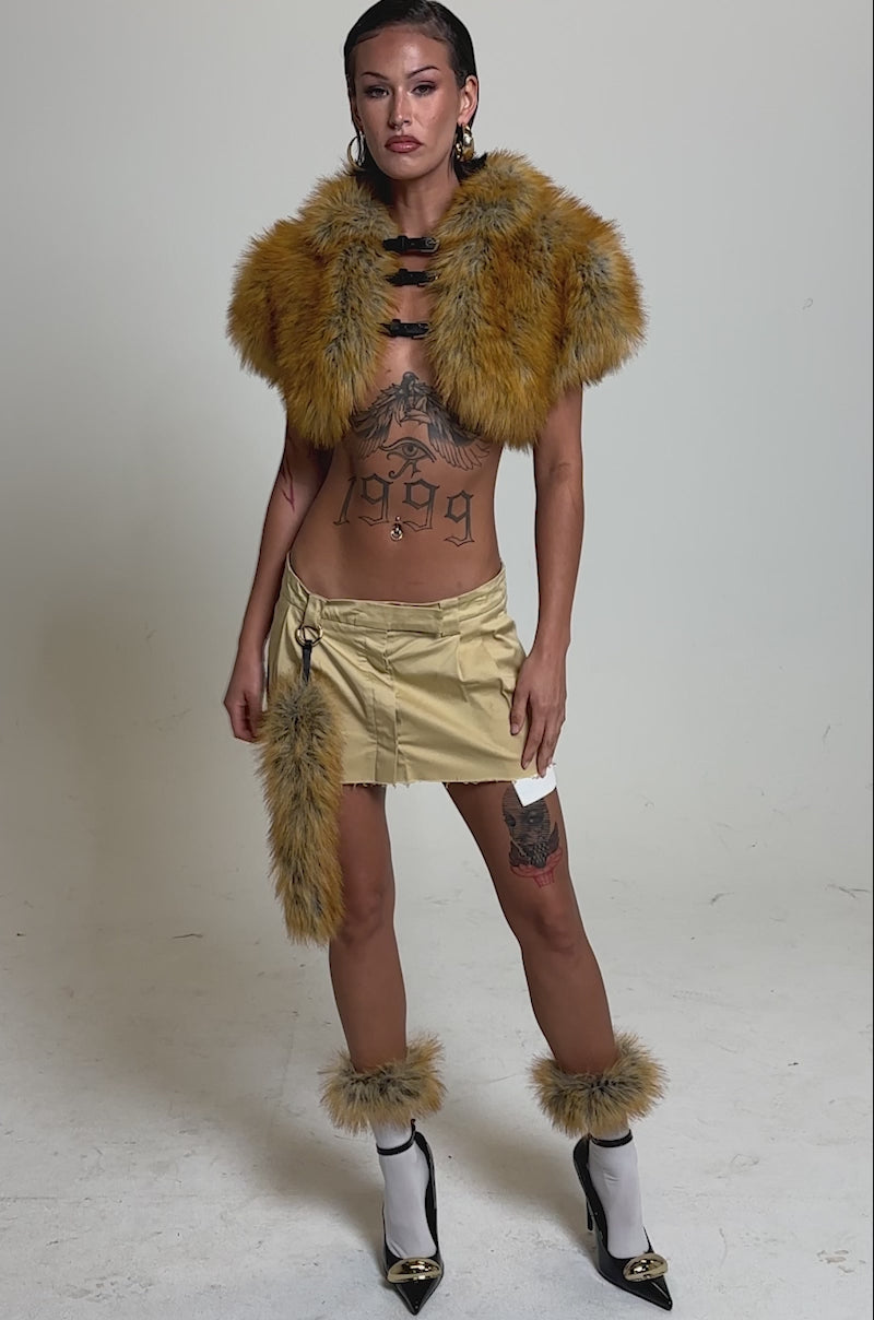 A video of a woman posing against a plain background wearing the LUXE LIFE FAUX FUR SHAWL IN BROWN, paired with a tan mini skirt featuring a faux fox fur tail, white socks with heels, and showcasing tattoos on her torso and legs.