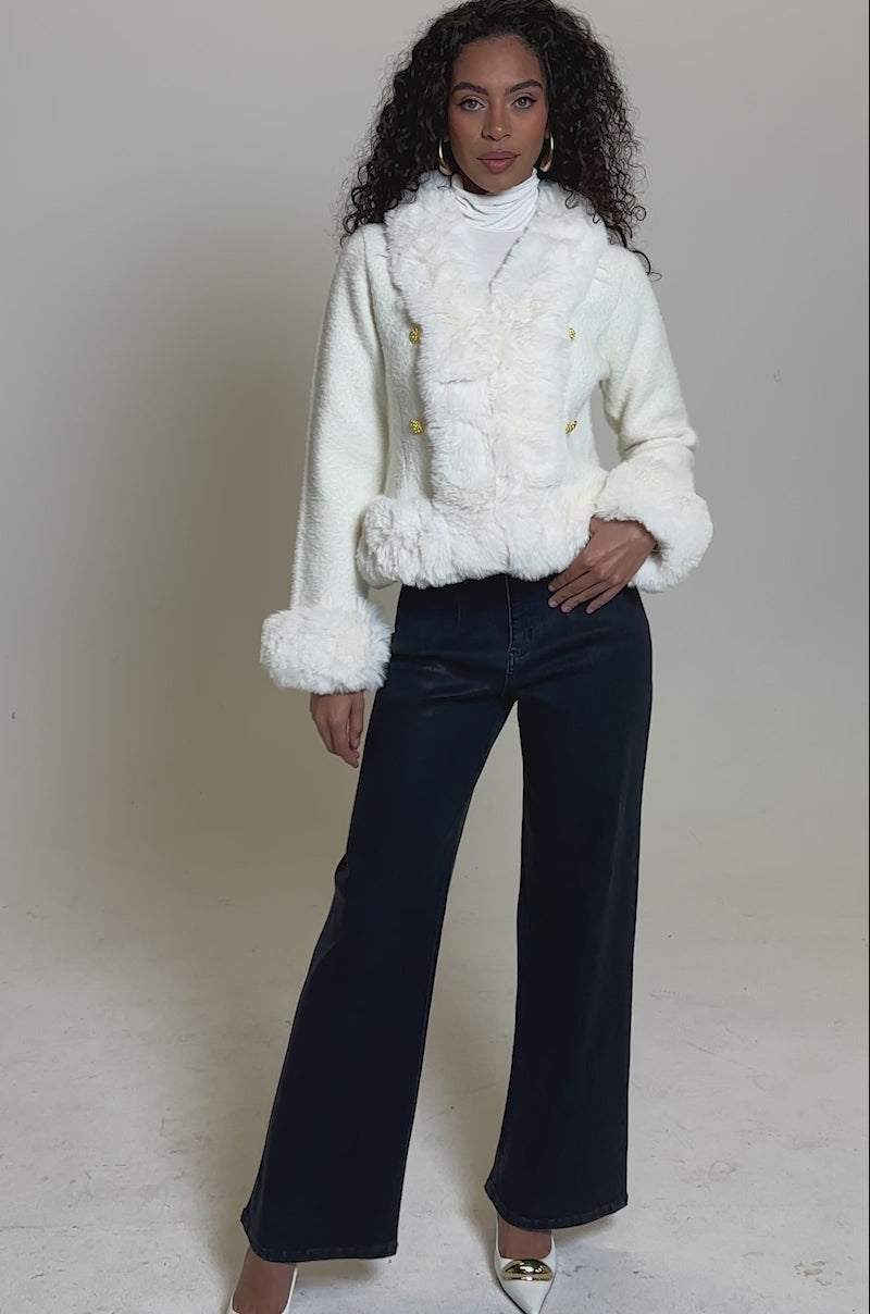 Video of woman modeling the SOFT TOUCH CARDIGAN, a white faux fur jacket with gold buttons, paired with a white top and dark blue high-waisted pants against a plain light background.