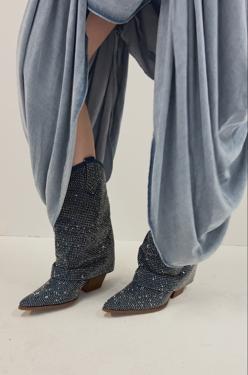 Video of person modeling the AZALEA WANG VANADIS DENIM RHINESTONE COWBOY BOOTIE features blue denim with rhinestones and small silver studs, a slouchy shaft, and a low, angled wooden heel.
