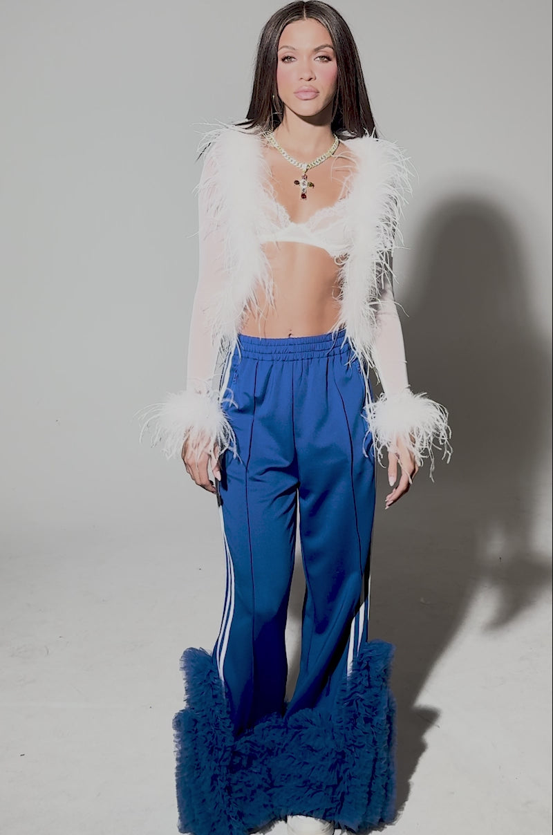 Video of woman modeling the MODERN JAM TRACK PANT WITH TULLE IN ROYAL BLUE—flared, with white stripes and large tulle ruffles at the cuffs—paired with white sneakers and a sheer top with feathery cuffs.