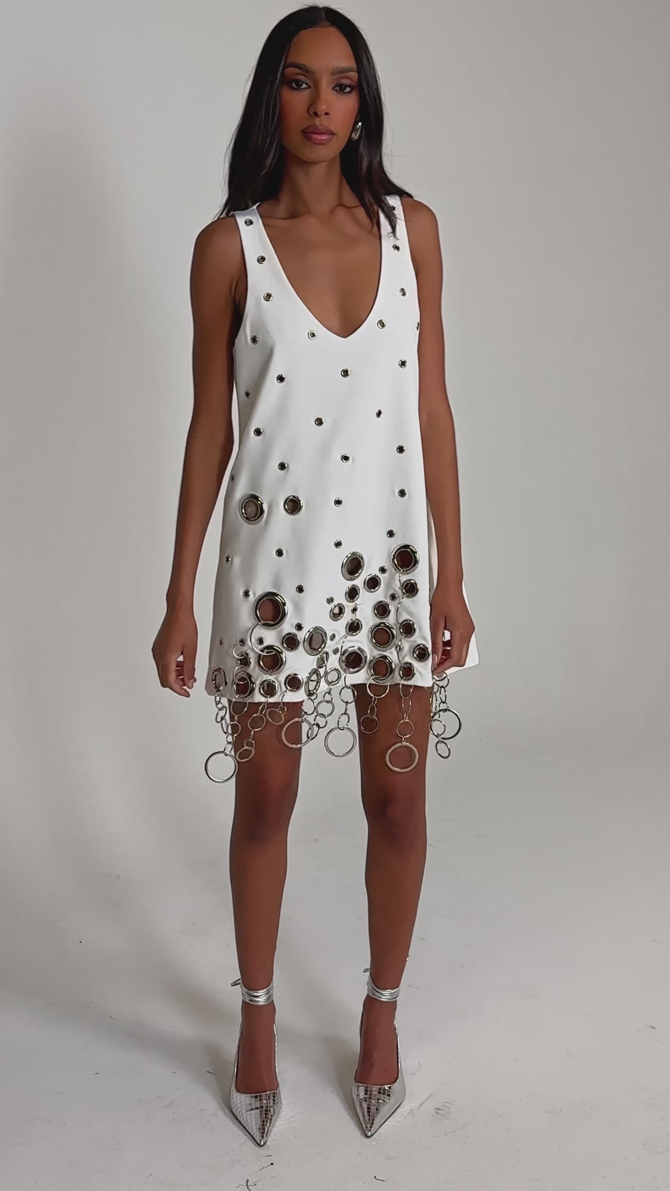 Video of woman modeling the ONE OF A KIND MINI DRESS WITH METAL HARDWARE IN WHITE, featuring a deep V-neckline, metallic eyelets, and large metal rings with chain fringe along the hem, set against a plain background.