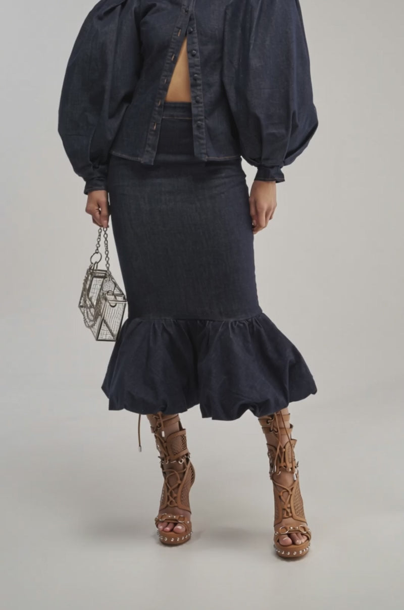 A video of a person wearing the LET'S CHANGE IT UP DENIM MIDI SKIRT—a high-waisted dark denim midi with a flared ruffle hem—paired with brown lace-up heeled sandals and a small geometric metallic handbag. 