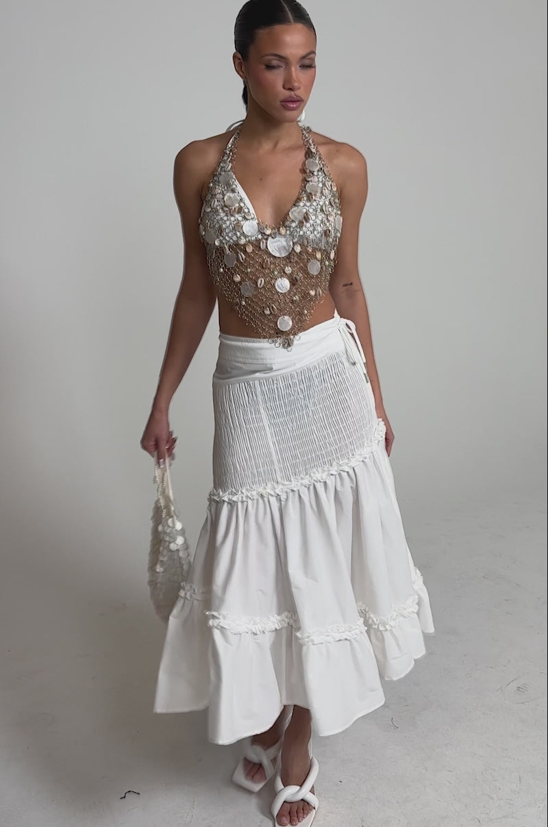 Video of woman modeling the THE BEST RUCHED SKIRT, a long, tiered midi skirt with ruffle trim and high-rise waist, paired with a sleeveless embellished top, white heeled sandals, and her hair pulled back. The background is plain and light.