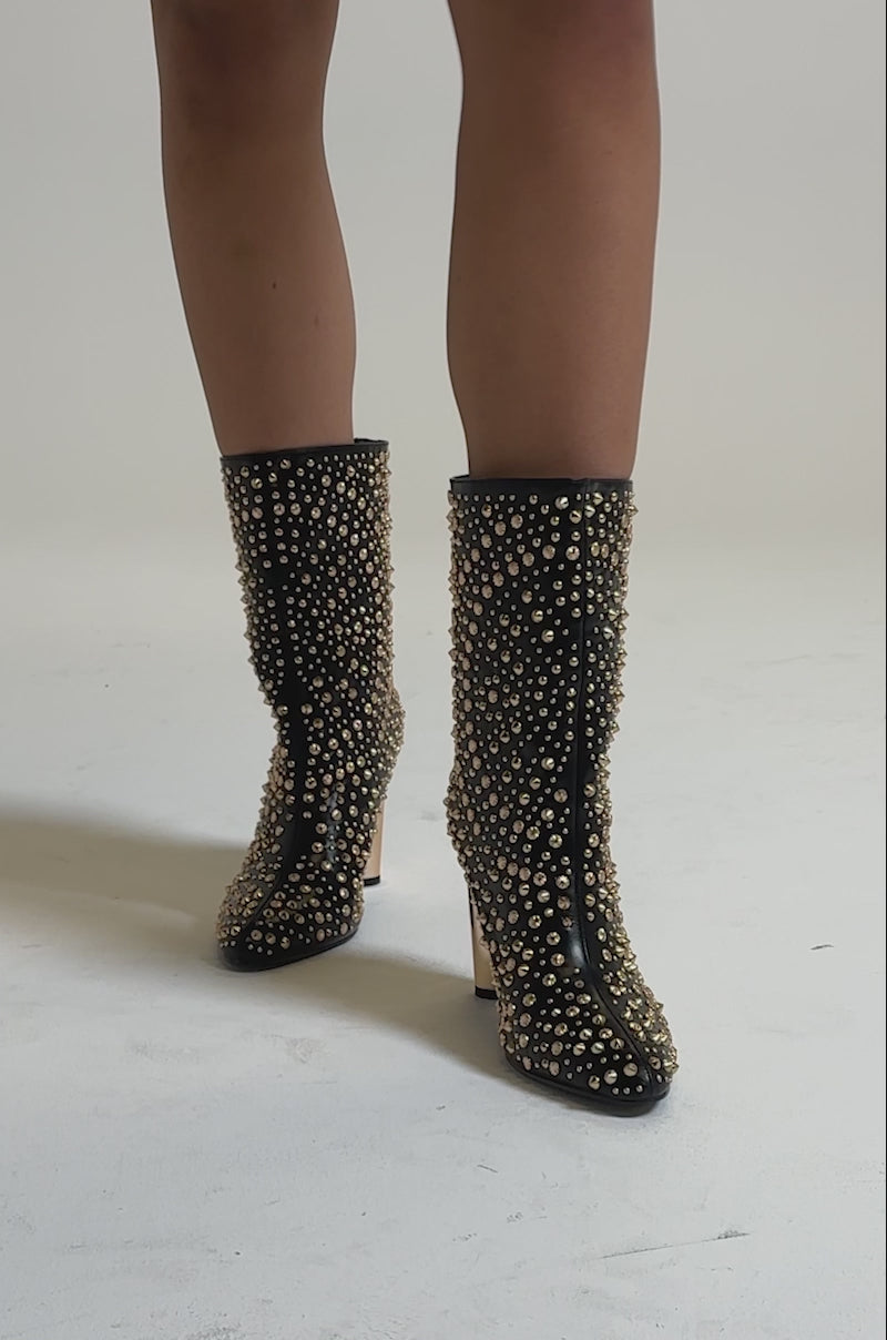 A video of the AZALEA WANG GREHGAN BLACK EMBELLISHED CHUNKY HEEL BOOTIE featuring black uppers, gold stud embellishments, pointed toes, and metallic gold block heels, seen worn by a person standing on a light surface.
