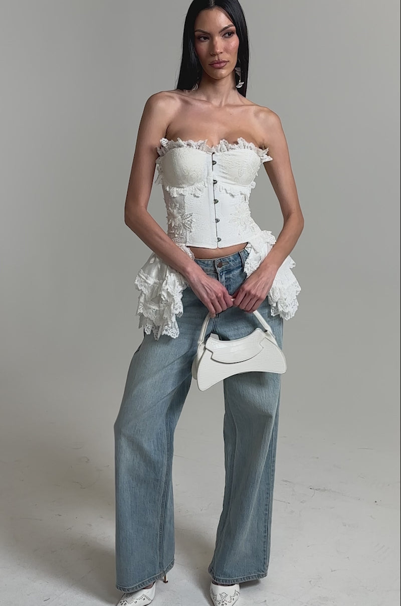 A video of a woman wearing the ALL WE DO LACE CORSET, styled with light blue wide-leg jeans, white pointed-toe heels, and a small white handbag.