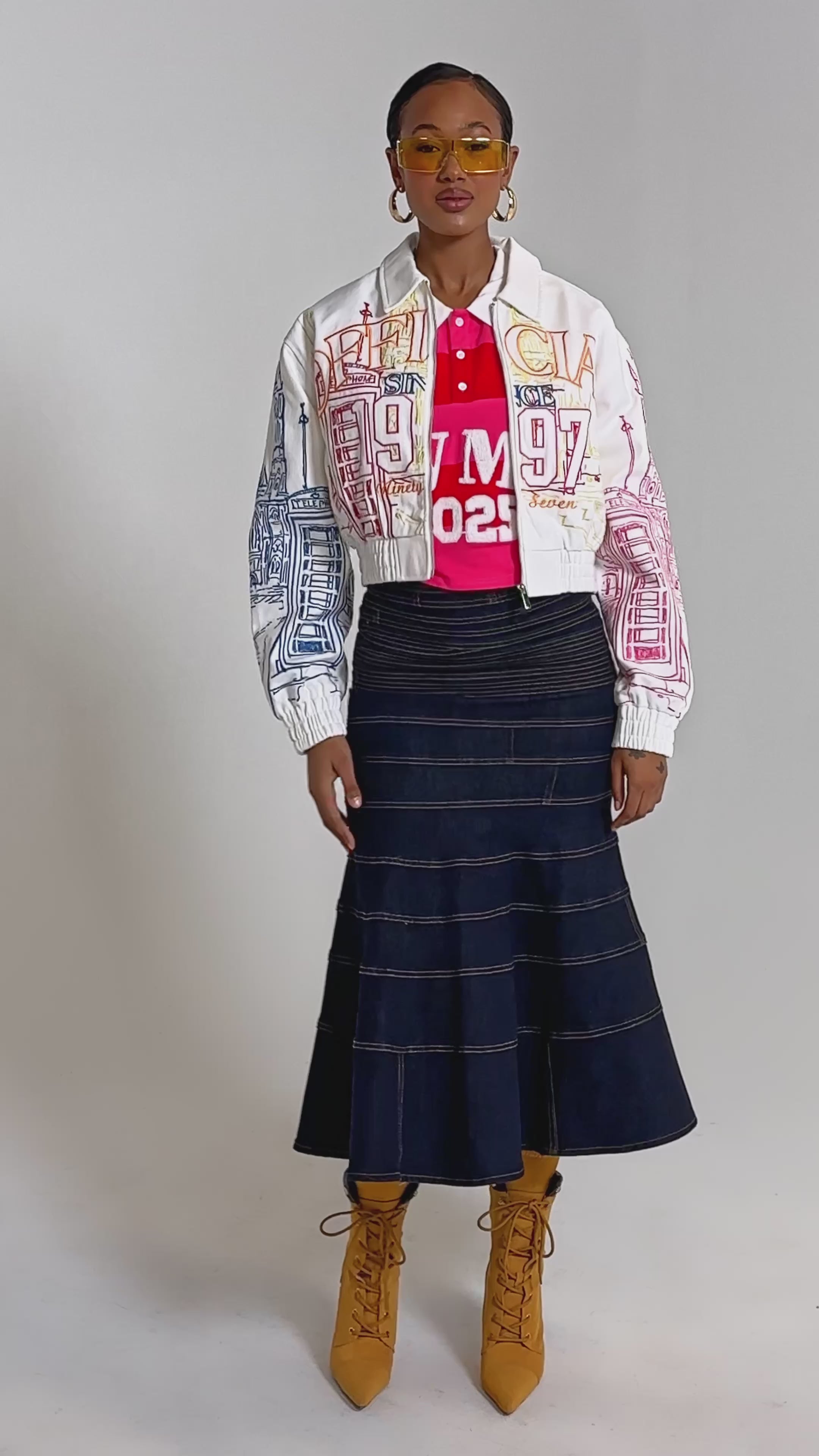 A video of a woman in the BEEN AROUND THE BLOCK EMBROIDERED BOMBER, styled with a red collared shirt, dark flared denim skirt, yellow-tinted sunglasses, and tan lace-up heeled boots.