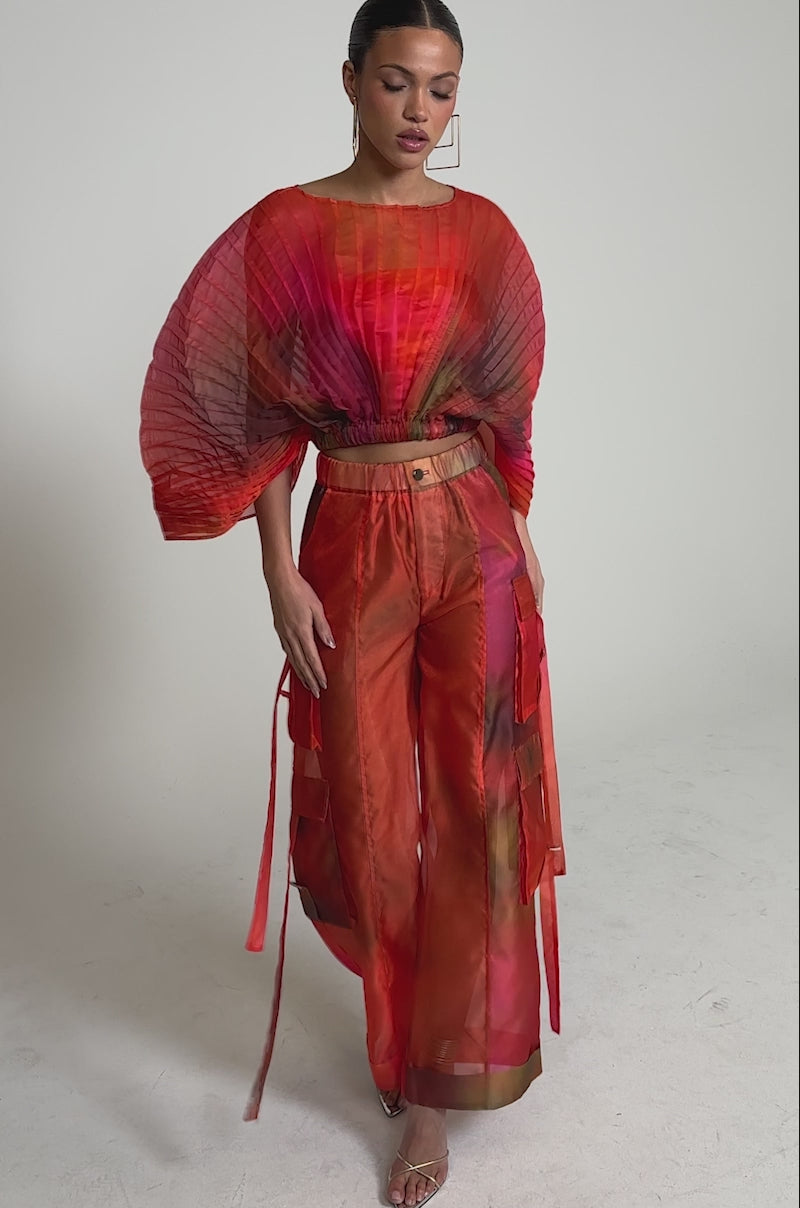 Video of woman modeling the STAINED RED CROPPED BLOUSE with dramatic, fan-like sleeves and matching high-waisted cargo pants, standing with one hand in her pocket against a plain background.