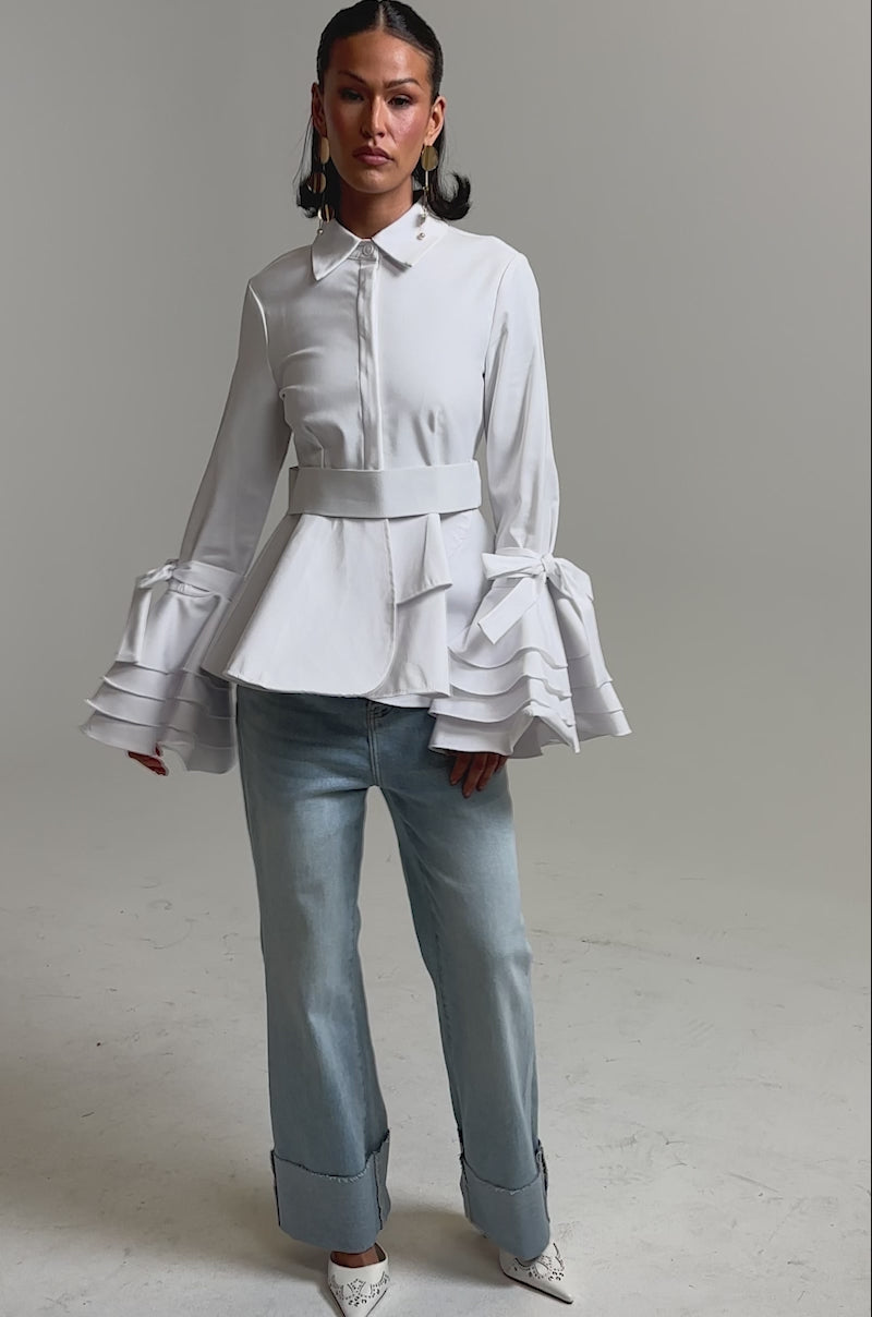 A video of a woman in the ELEGANT PEPLUM BUTTON DOWN BLOUSE with tiered ruffle cuffs, styled with light blue cuffed jeans and white pointed heels against a plain light background.