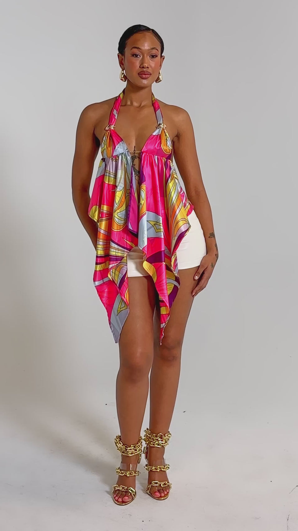 A video of a woman in the FUN AND FLIRTY SATIN BLOUSE with an asymmetrical hem, cream shorts, gold chain-detail heeled sandals, and a leopard-print clutch.