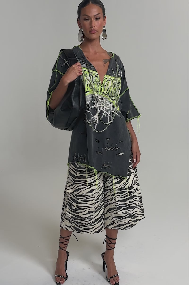 Video of woman modeling the KNOCKOUT OVERSIZED TEE, featuring distressed details, neon green stitching, safety pins, and bold “KNOCKOUT” text. She completes her streetwear look with zebra-print pants, hoop earrings, and a black bag.