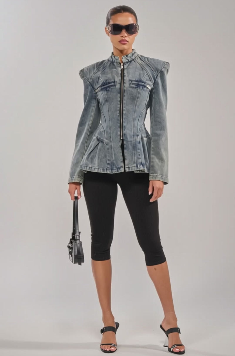 A video of a woman wearing the MALLORCA PEPLUM DENIM JACKET, black cropped leggings, black heeled sandals, sunglasses, and holding a small black handbag stands against a plain light background.