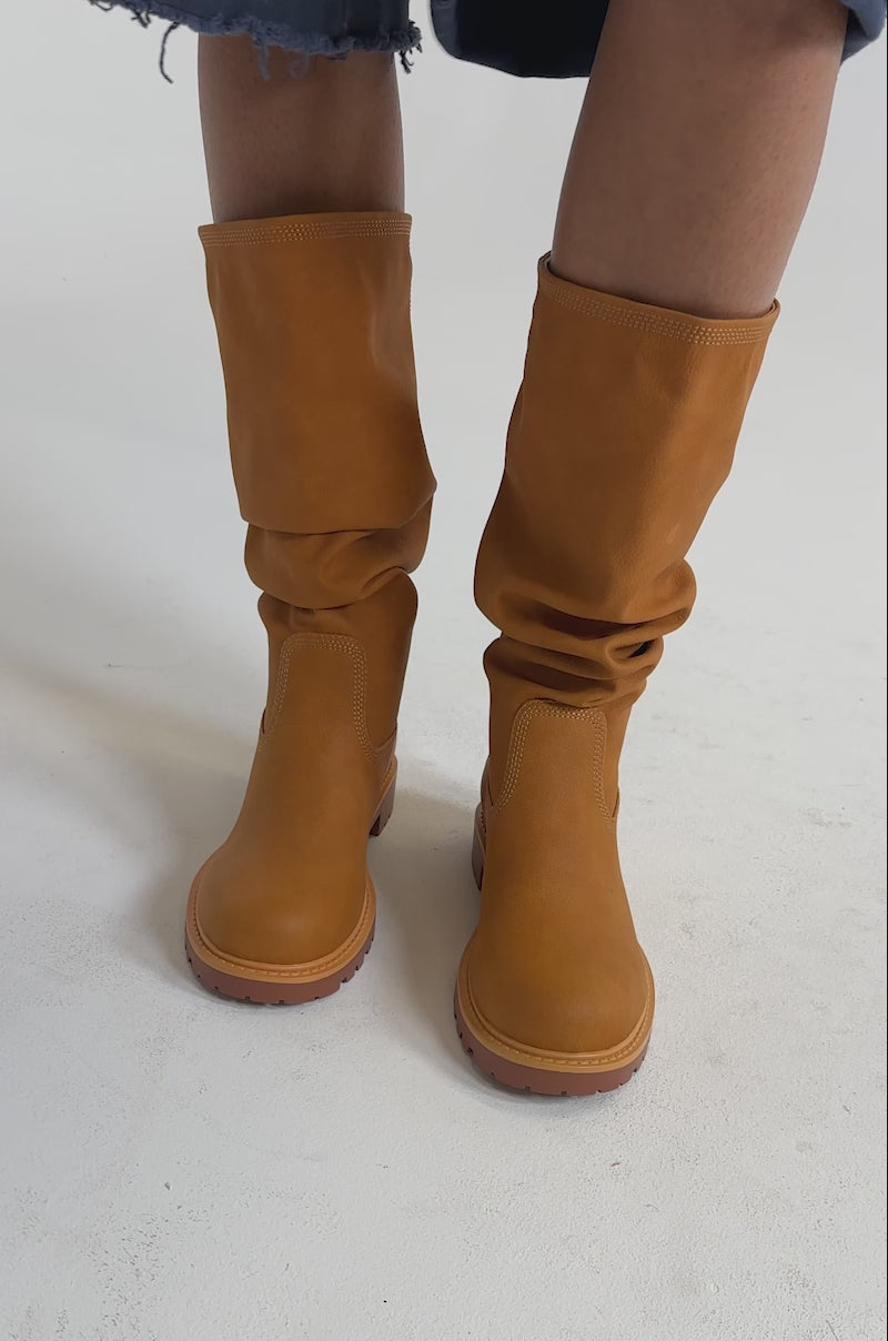 A video of a person wearing the AKIRA GOLD BARTLETT TAN FLAT RUCHED BOOTIE and knee-length blue denim cargo shorts with frayed hems stands against a light background. 