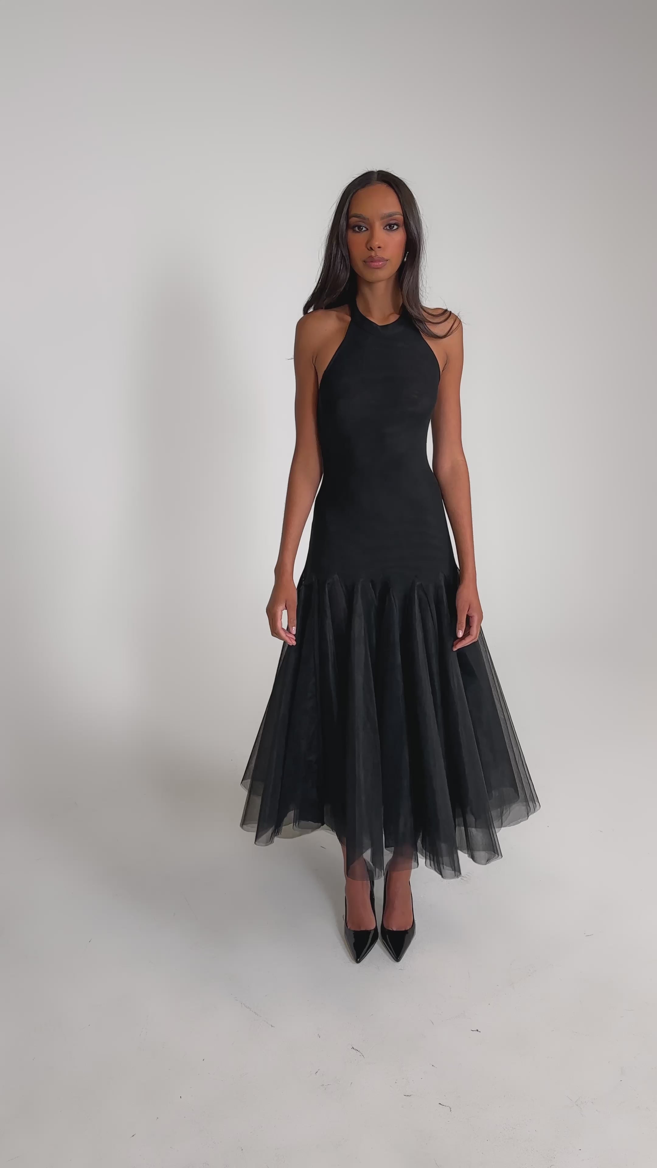 Video of woman modeling the SWEPT UP MESH MAXI DRESS, a black halter style with a tulle mermaid skirt, paired with black heels. She has long hair and stands against a plain light gray background.