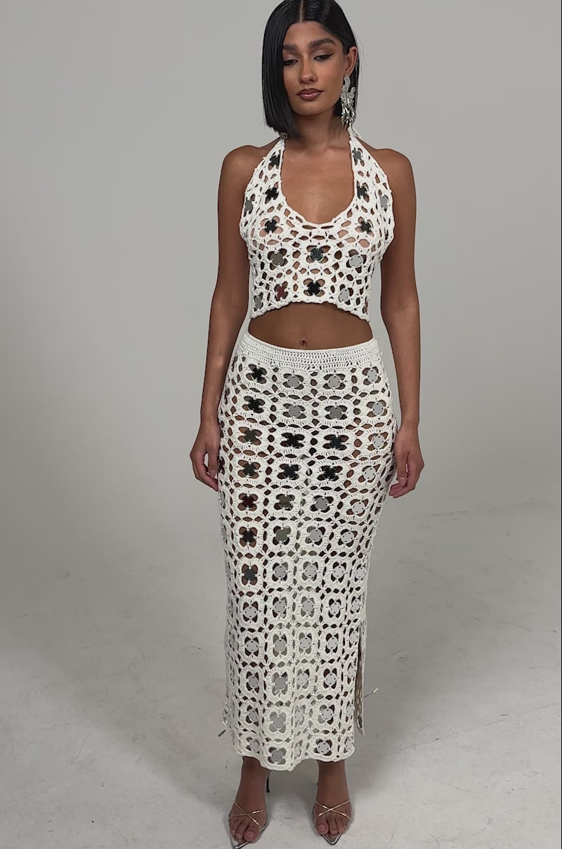 A video of a woman wearing the LETS GO TO THE RESORT MAXI SKIRT, a cream crochet maxi with a slit and multicolored florals, paired with a matching halter-neck crop top and heels.