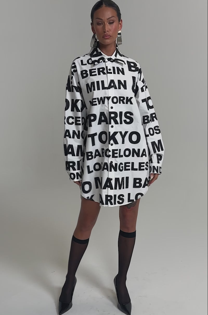 Video of woman modeling the TIME AND PLACE POPLIN MINI DRESS, a black and white long-sleeved mini shirt dress featuring bold city names like Milan, Tokyo, Paris, and New York, against a plain gray background.