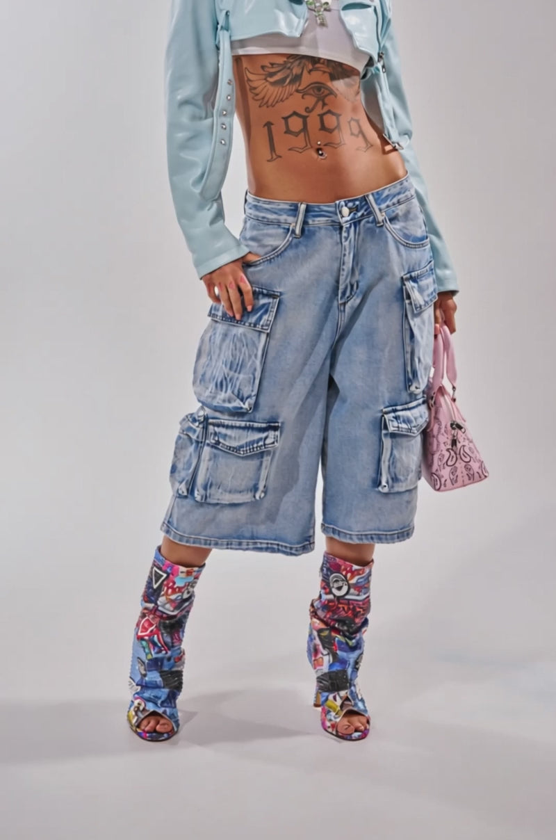Video of woman modeling the TOO MUCH FUN CARGO BERMUDA SHORTS, a light blue cropped jacket, sheer crop top, graphic boots, layered necklaces, and a pink handbag—an eye-catching festival look.