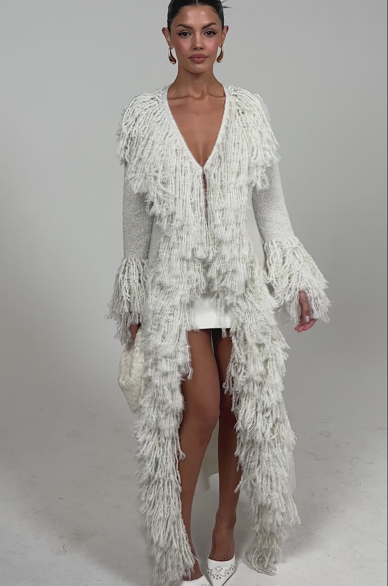 Video of woman modeling the BURRR IT'S COLD OUTSIDE FRINGE OVERSIZED SWEATER IN IVORY over a white mini dress, styled with white pointed heels, gold hoop earrings, and a woven white handbag against a plain light background.