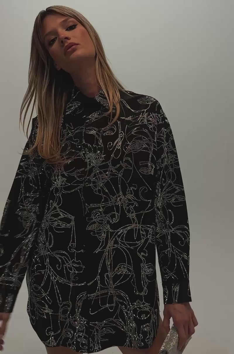 Video of a woman modeling the ZIG AND ZAG POPLIN MINI DRESS—a long-sleeved black mini dress with a white abstract line pattern and button-up front. 