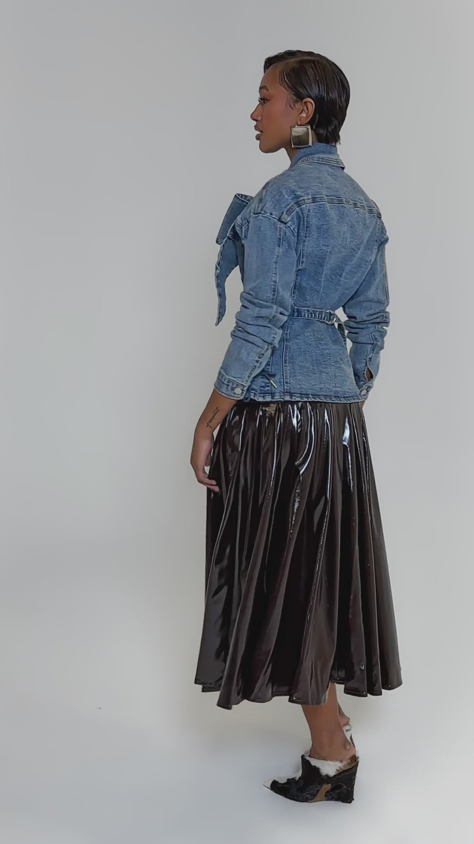 Video of woman modeling the DAY BREAK BUTTON DOWN BLOUSE, paired with large silver earrings and a black shiny skirt, stands facing forward against a plain light background.